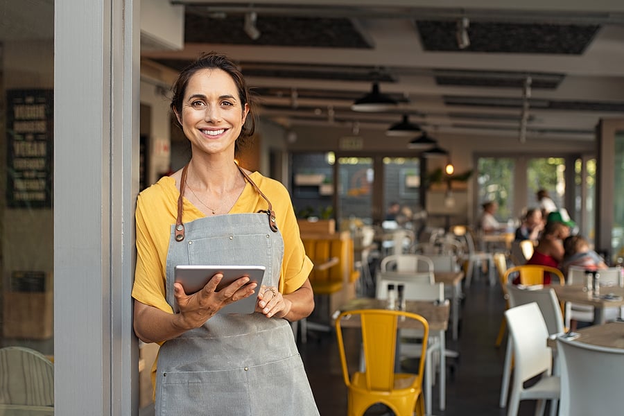 A smiling café server wearing an apron holds a tablet while standing at the entrance of a bright, modern restaurant with seated diners and yellow chairs in the background.